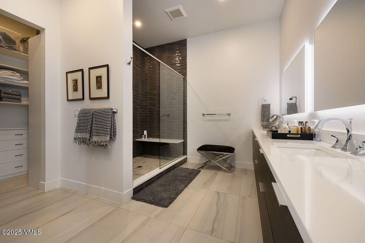 This is a well-lit primary bathroom featuring a modern design with a walk-in shower enclosed by glass, a double vanity with white countertops and dark wood cabinets, and neutral-toned tile flooring. The space is decorated with framed artwork, towels, and a small bench, creating a clean and luxurious atmosphere. A walk-in closet is visible to the left.
