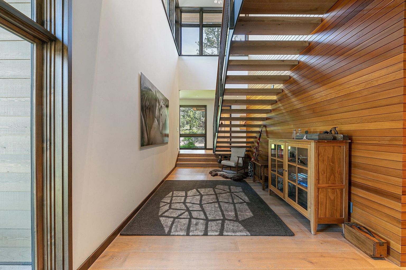 This interior shot showcases a modern hallway with wooden stairs and paneling. A large rug with a geometric pattern lies on the wooden floor, leading towards a doorway with natural light streaming in. A cabinet sits against the wood-paneled wall, adding a touch of elegance and functionality to the space.