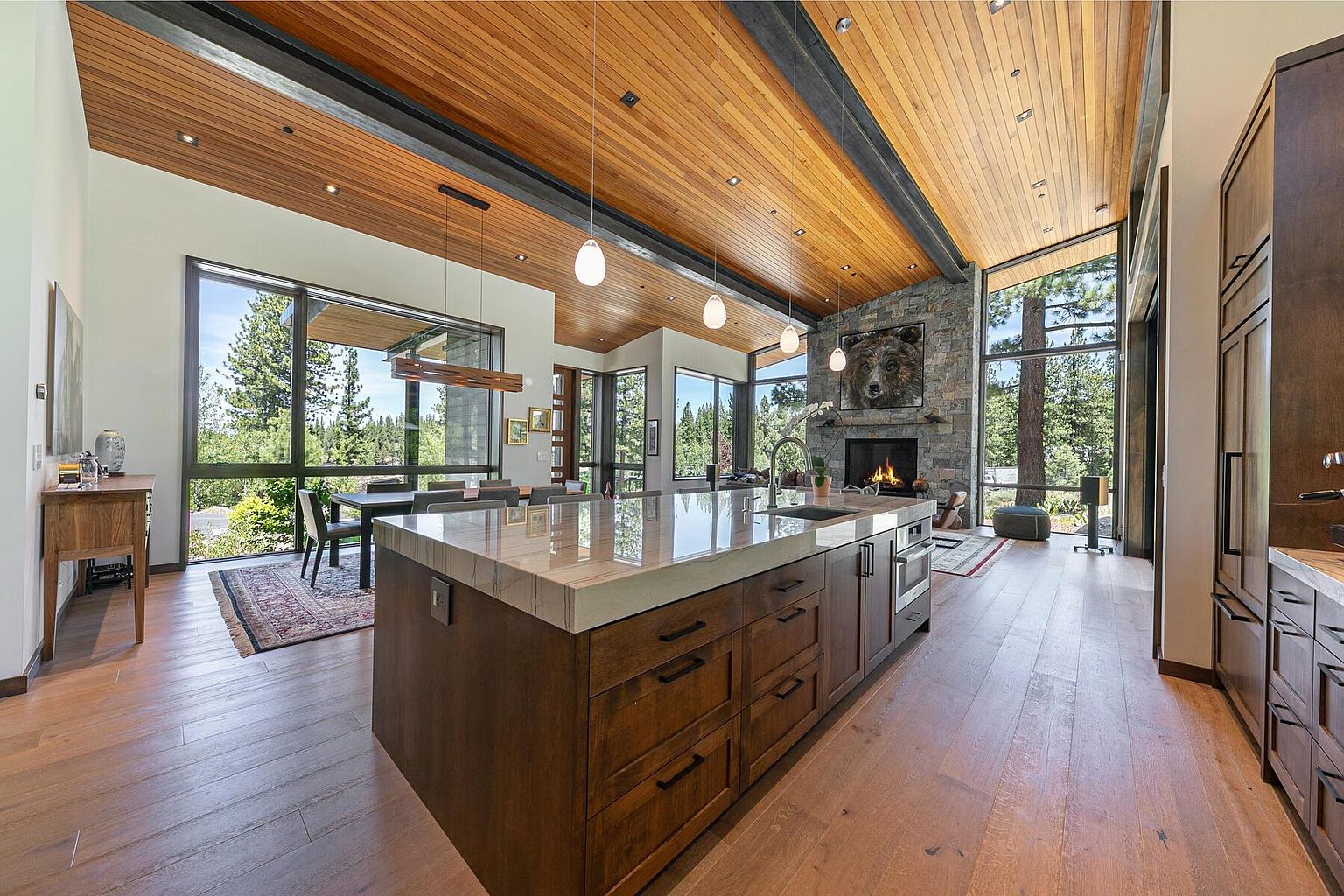 This is a wide-angle shot of a modern, luxurious kitchen featuring a large island with a marble countertop and dark wood cabinetry. The kitchen boasts high ceilings with wood paneling and exposed steel beams, complemented by pendant lighting. Large windows offer expansive views of the surrounding trees, and a stone fireplace with a bear artwork adds a rustic touch to the contemporary design.