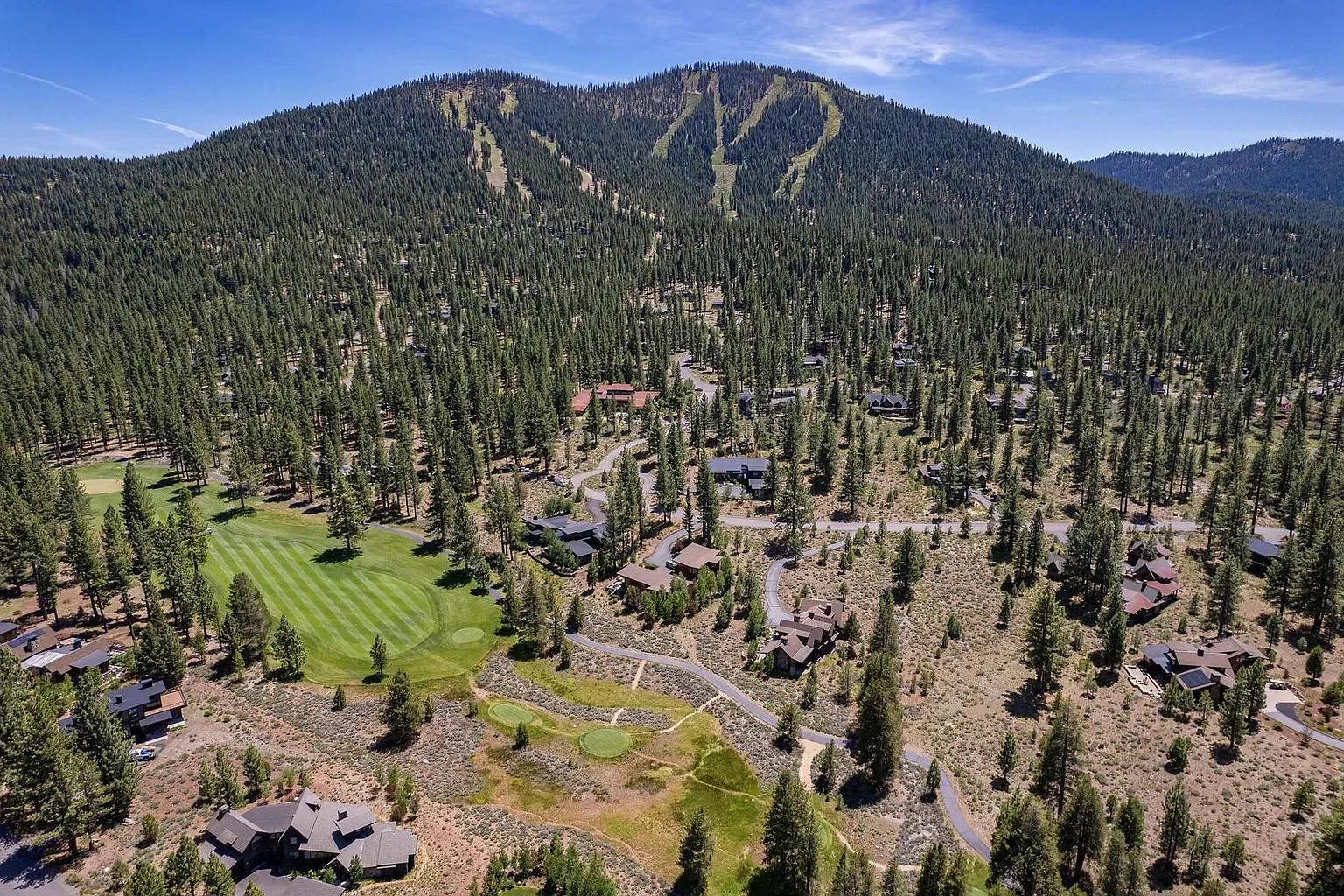 This aerial view showcases a high end mountain community. Several luxury homes are nestled among lush green trees and a well-maintained golf course. Ski slopes are seen carved into the mountain in the background.
