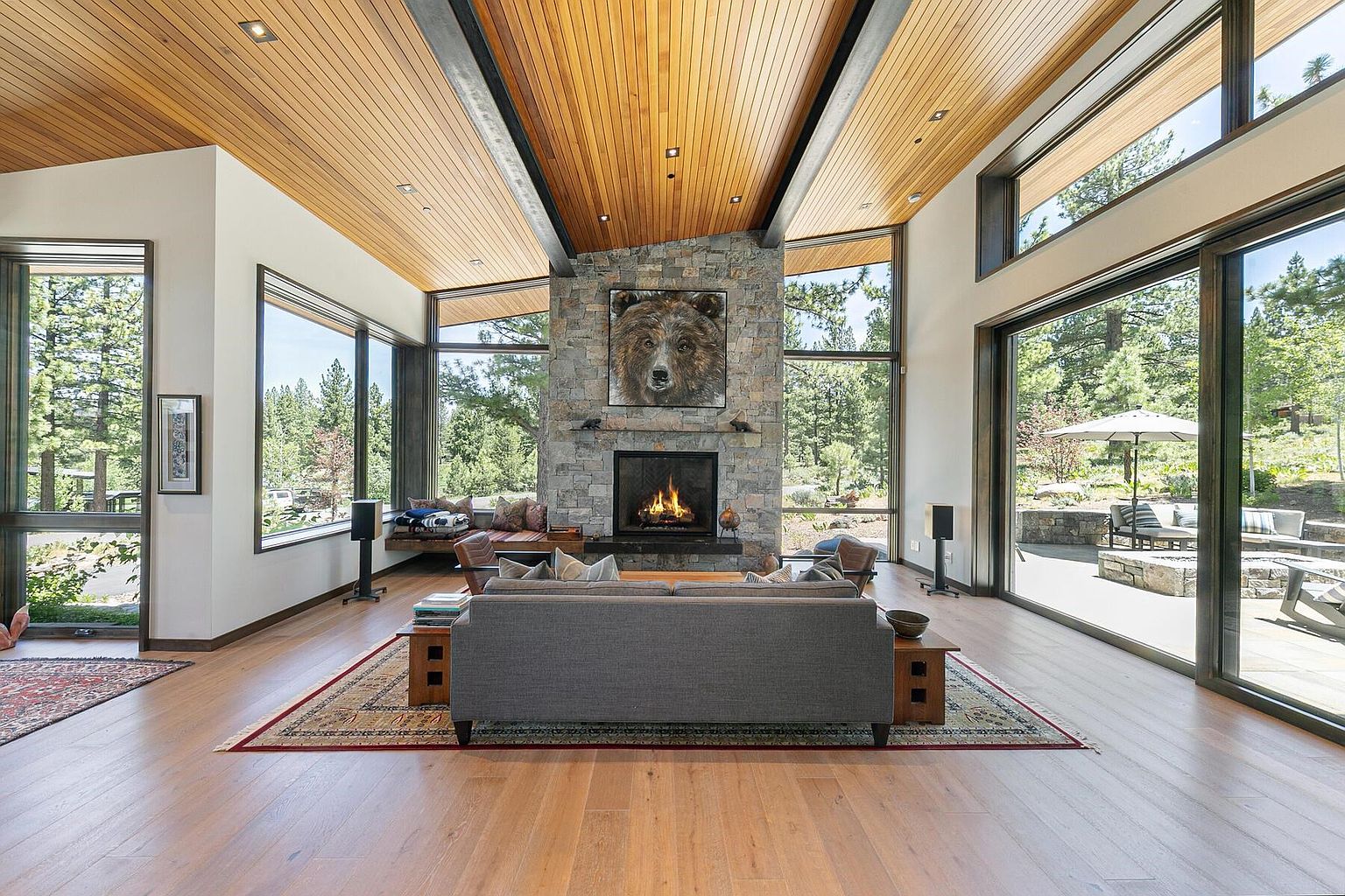 This is a spacious living room featuring a stone fireplace with a bear portrait above it, large windows offering views of the surrounding trees, and a wood-paneled ceiling. A gray sofa sits on a patterned rug, and the room is filled with natural light. The overall impression is one of modern luxury and connection to nature.