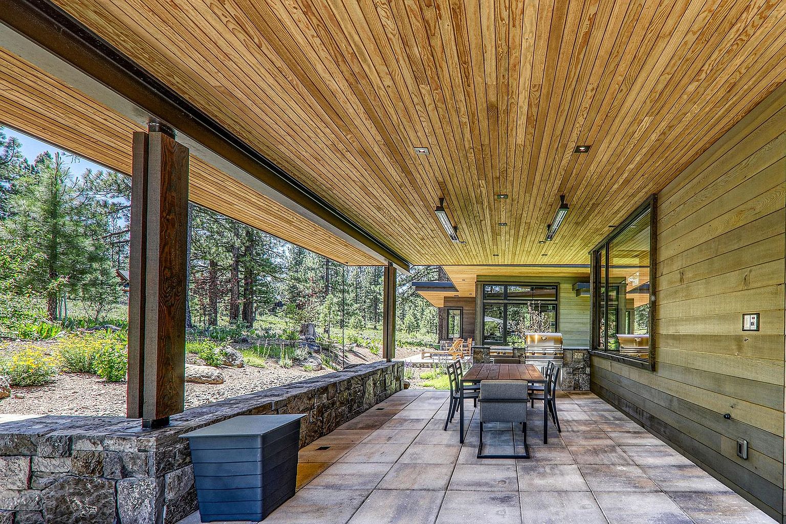 This image showcases a beautifully designed outdoor patio area, featuring a wooden ceiling, stone accents, and a dining set. The patio offers a seamless transition between indoor and outdoor living, with views of a lush, natural landscape. The space is well-lit and appears perfect for entertaining or relaxing.