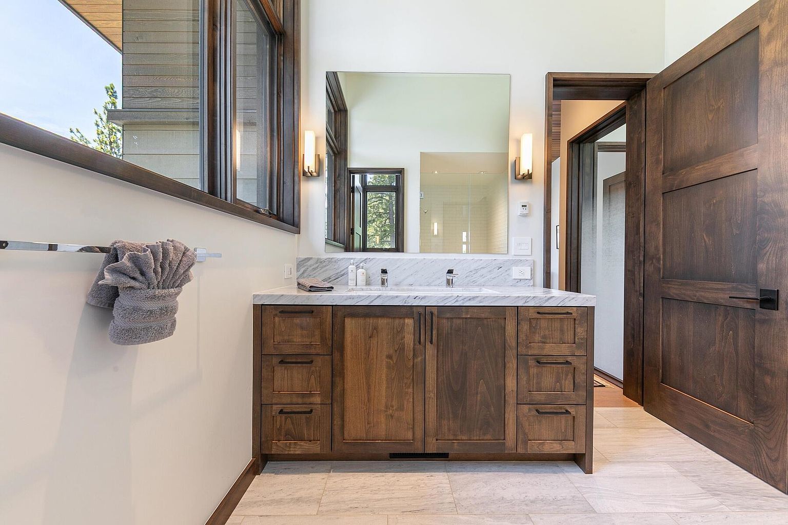 This is a well-lit primary bathroom featuring a double vanity with a marble countertop and dark wood cabinetry. A large mirror hangs above the vanity, flanked by sconce lighting. The room has a modern, clean aesthetic with neutral-toned tile flooring and a glimpse of a shower through an open doorway.