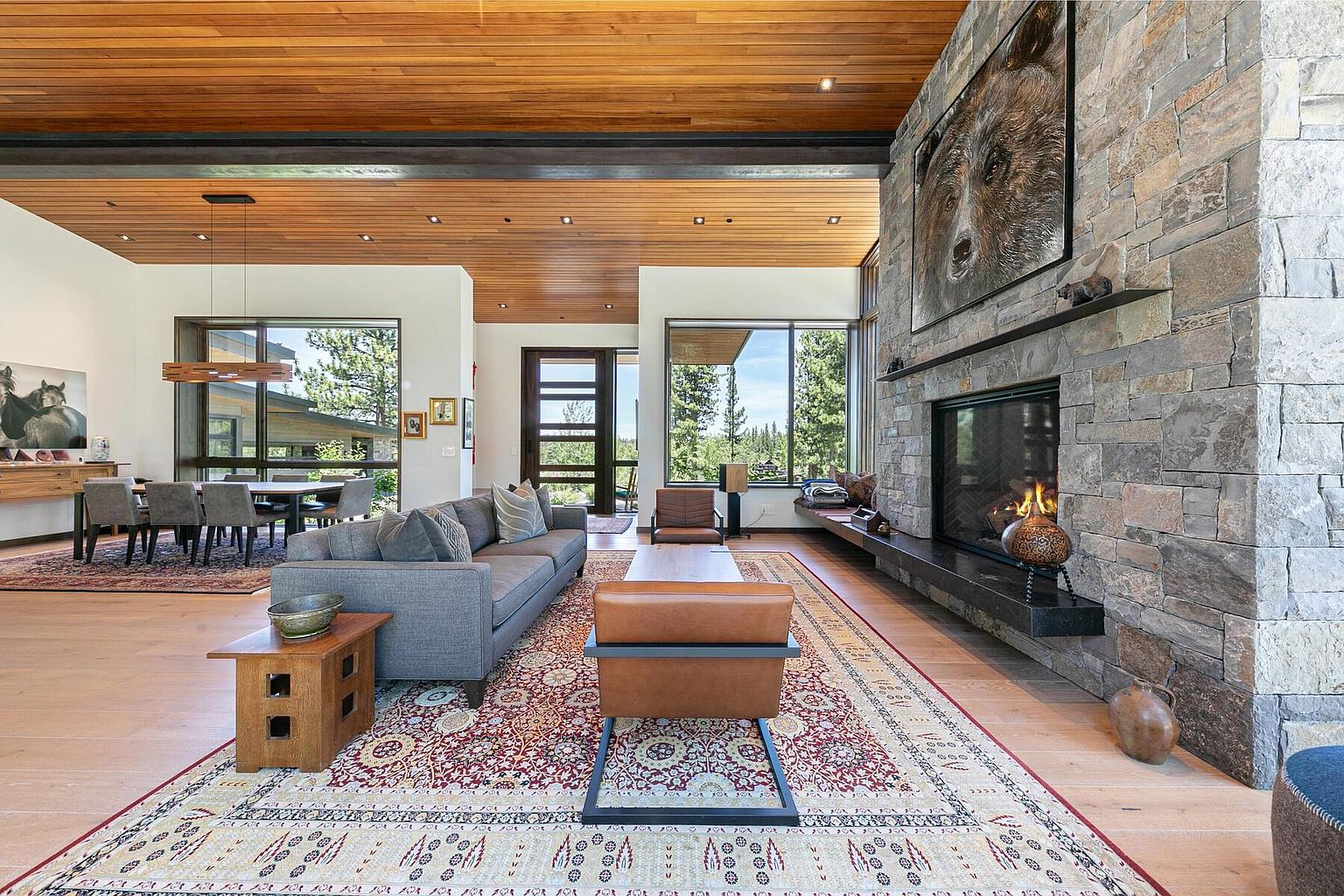 This is a spacious living room featuring a stone fireplace with a large bear artwork above, a gray sofa, and a patterned rug. The room has large windows offering natural light and views of the outdoors. The wooden ceiling adds warmth to the modern design.