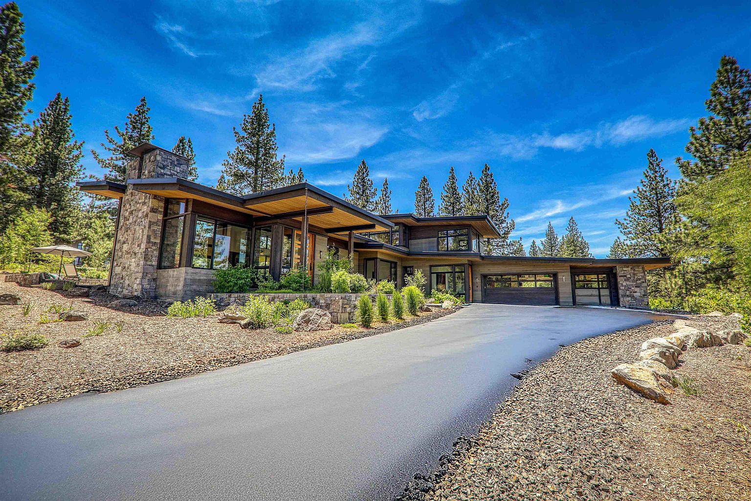 This is a front exterior view of a modern, luxurious home. The house features a combination of stone and wood elements, large windows, and a sleek, low-slung roofline. A paved driveway leads up to the house, surrounded by landscaping with rocks and greenery, creating a sophisticated and inviting curb appeal.