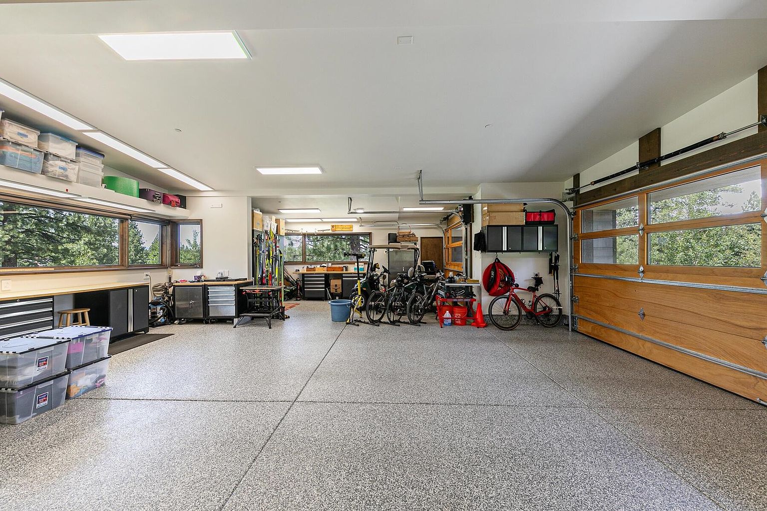 This is an interior shot of a well-organized garage. The garage features a speckled epoxy floor, a large wooden garage door with windows, and ample storage solutions including cabinets, shelving, and workbenches. Bicycles, tools, and storage bins are neatly arranged, suggesting a functional and tidy space.