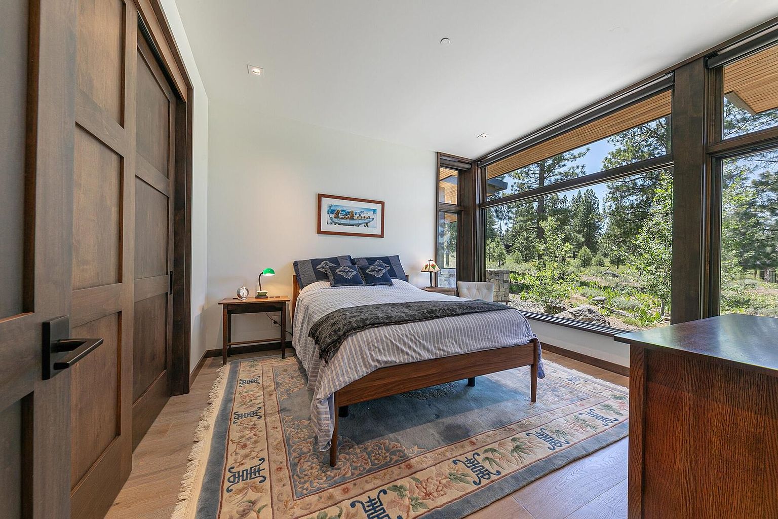 This is a cozy bedroom featuring a wooden bed frame with striped bedding and decorative pillows, complemented by a patterned area rug. A large window offers a view of the surrounding greenery, enhancing the room's natural light and serene atmosphere. The room also includes a wooden side table with a lamp and a wooden dresser, adding to the room's warmth and functionality.