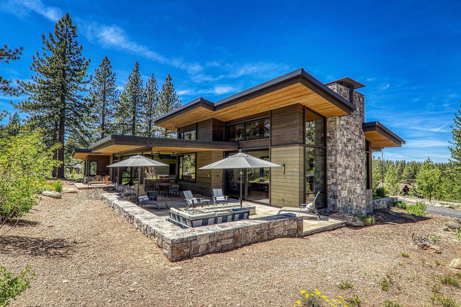 This image showcases the rear exterior of a modern home, highlighting a spacious patio area with a fire pit and outdoor seating. The architectural design features a combination of wood and stone elements, with large windows offering ample natural light. The surrounding landscape includes mature trees and a well-maintained yard, creating a serene and inviting atmosphere.