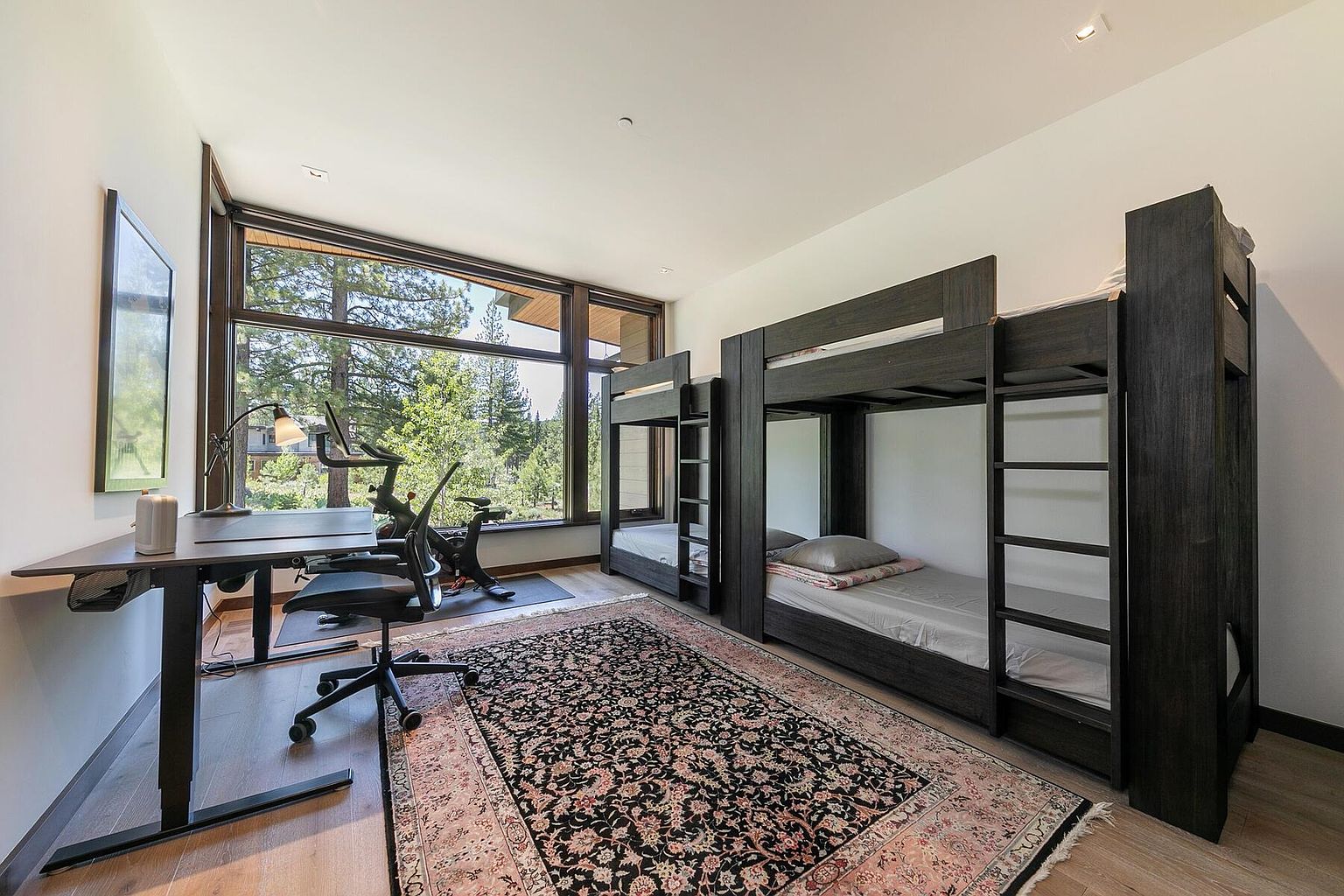 This bedroom features a modern design with dark wood bunk beds and a large window offering a view of the surrounding trees. A desk and exercise bike are positioned near the window, creating a versatile space for work and fitness. The room is grounded by a patterned area rug, adding warmth and character to the space.