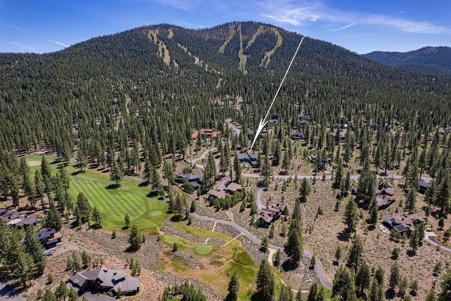 This aerial shot showcases a luxurious property nestled amidst a dense forest and a golf course. The home features a dark roof and is surrounded by mature trees, offering privacy and seclusion. In the background, a mountain with ski runs adds to the scenic beauty of the location.