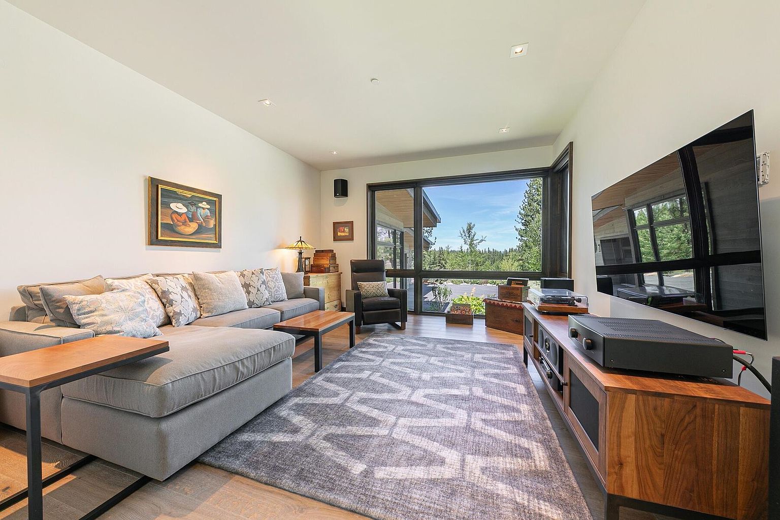 This is a well-lit living room featuring a large gray sectional sofa with decorative pillows, a modern area rug with a geometric pattern, and a wooden entertainment center with a large flat-screen TV. A large window provides natural light and a view of the outdoors. The room has a contemporary style with a focus on comfort and entertainment.