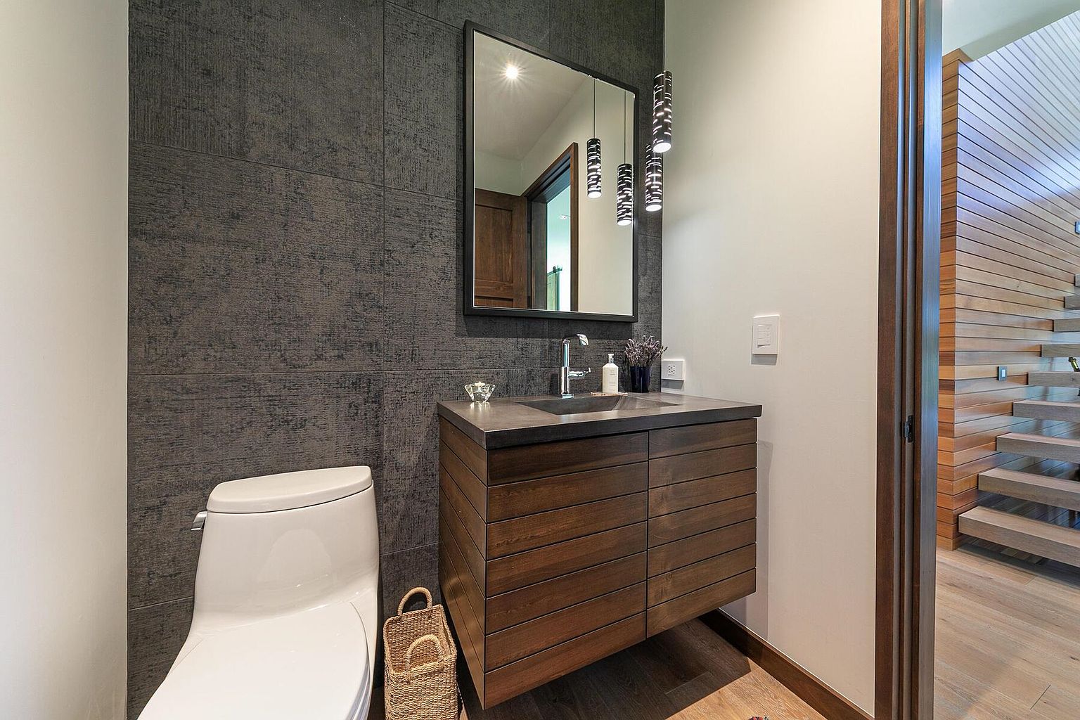 This is a modern guest bathroom featuring a floating wood vanity with a dark countertop and a rectangular mirror above. The wall behind the vanity is covered in textured gray tiles, contrasting with the white walls on the other side of the room. A toilet is visible to the left, and a doorway leads to a staircase with wooden steps and a matching wooden accent wall, creating a cohesive and stylish design.