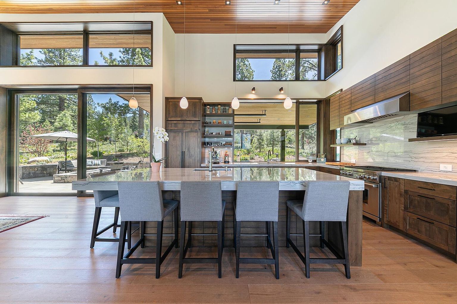 This is a bright and modern kitchen with a large island featuring seating for four. The kitchen boasts wooden cabinetry, stainless steel appliances, and large windows that offer a view of the outdoor landscape. The open floor plan and natural light create an inviting and spacious atmosphere.