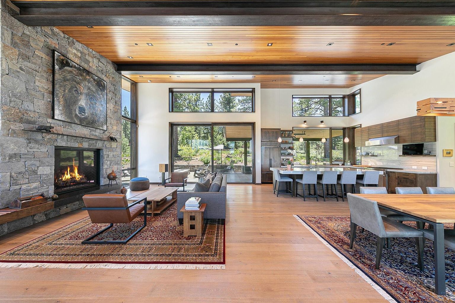 This is a spacious open-concept living area featuring a stone fireplace with a large bear artwork above it, a modern kitchen with an island and bar stools, and a dining area. The room is well-lit with natural light from large windows and features hardwood floors and exposed wooden beams. The overall impression is luxurious and inviting, perfect for entertaining.