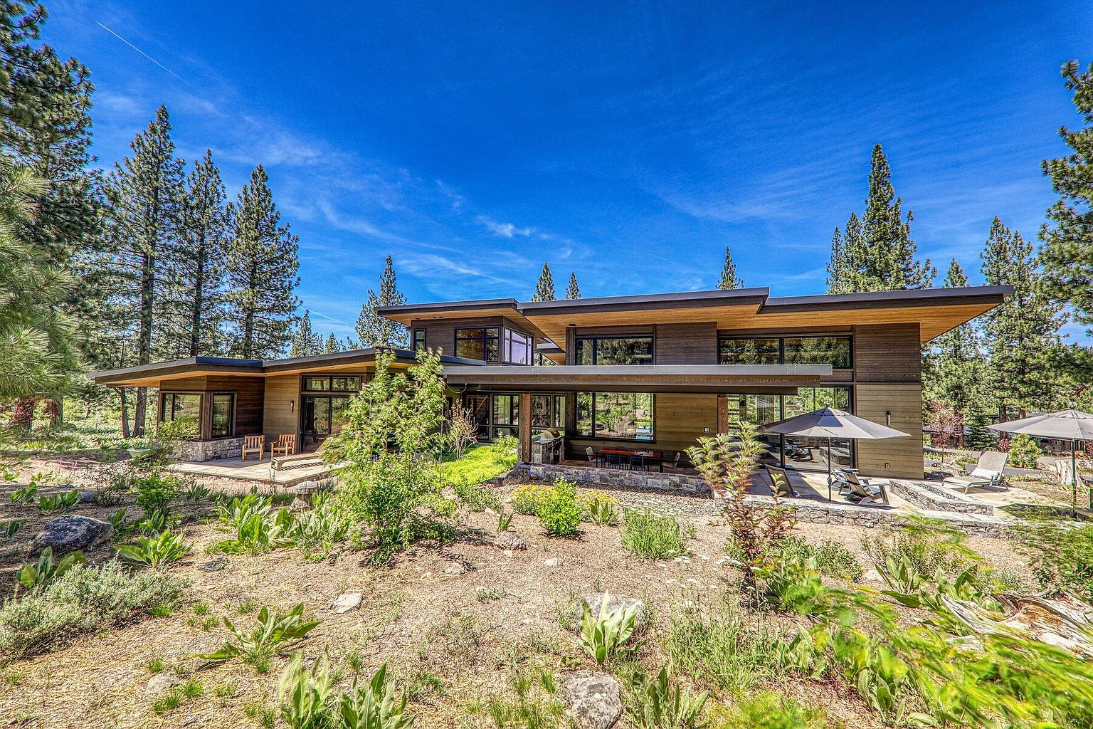 This image showcases the rear exterior of a modern home, emphasizing its integration with the natural landscape. The architecture features clean lines, wood siding, and expansive windows that offer views of the surrounding trees and sky. A patio area with outdoor seating and umbrellas extends from the house, creating an inviting space for relaxation and entertainment.