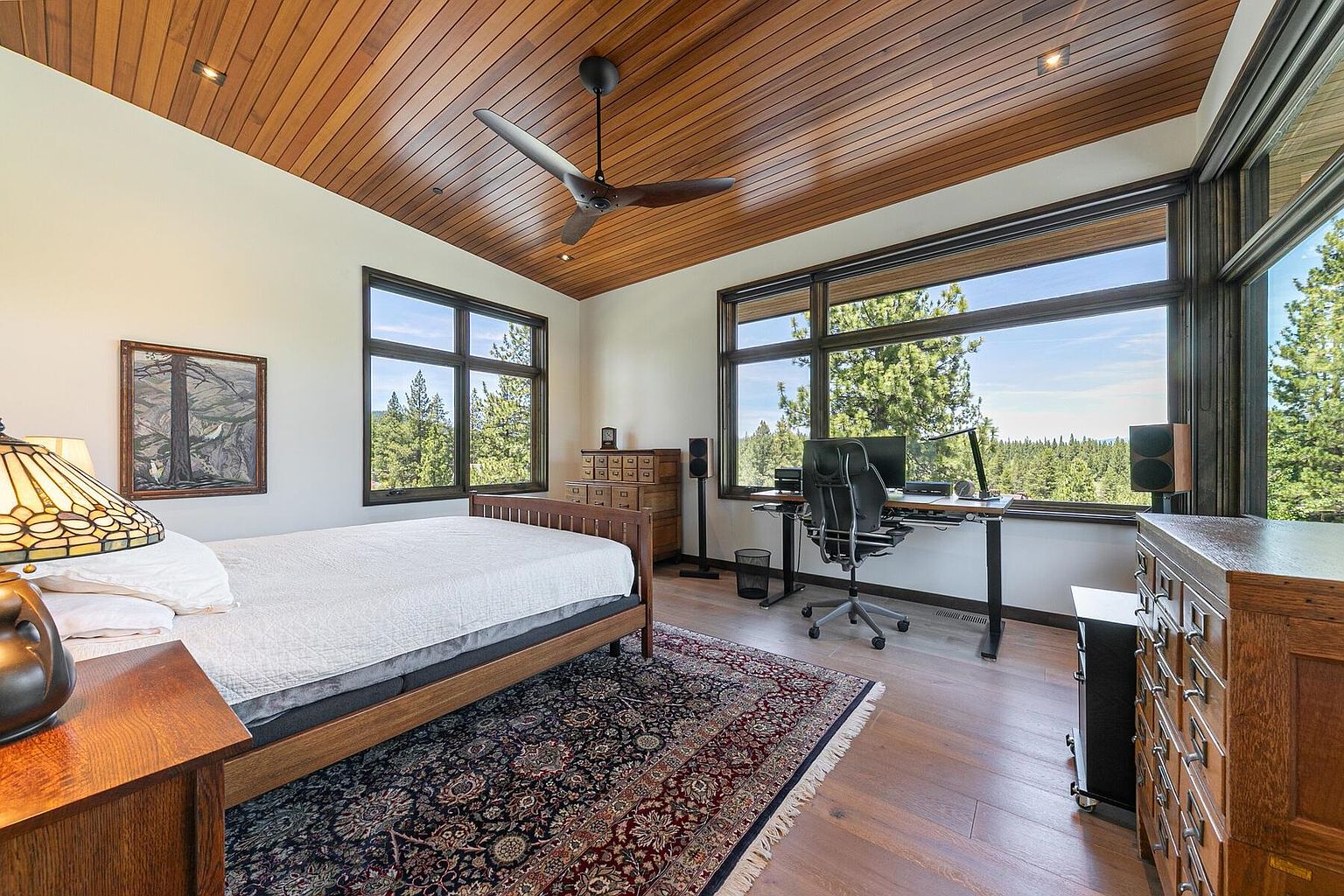This is a primary bedroom featuring a wooden ceiling and a large window with a view of the forest. The room includes a bed, a desk with a computer, and wooden dressers. The style is rustic and modern, creating a cozy and functional space.