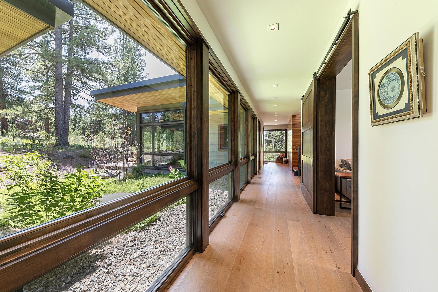 This interior shot showcases a modern hallway with wood flooring and large windows offering views of the exterior landscape. The hallway features wooden framed windows and a sliding barn door, adding a rustic touch to the contemporary design. An artwork hangs on the wall, enhancing the aesthetic appeal of the space.