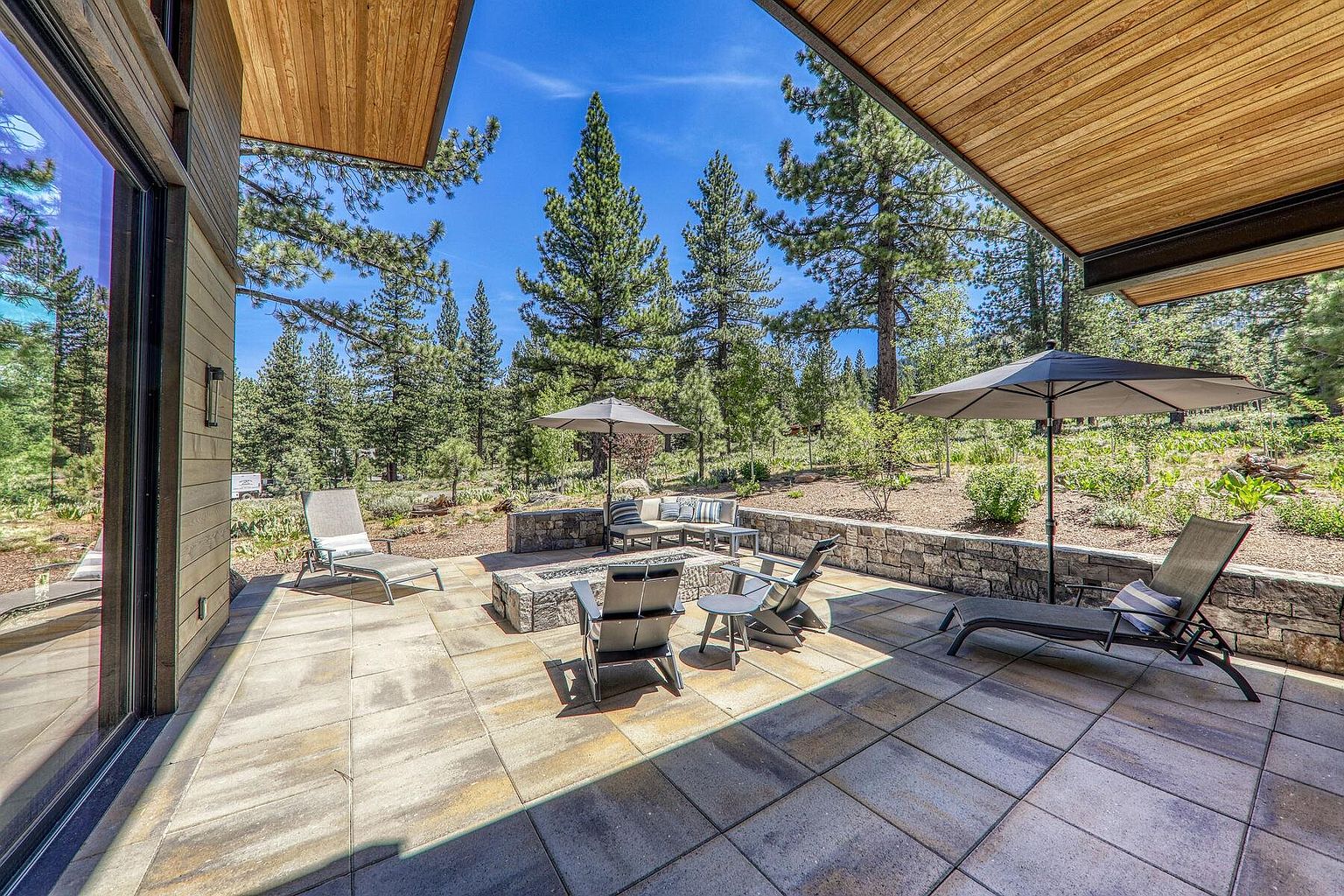 This image showcases a beautifully designed outdoor patio area, perfect for relaxation and entertainment. The patio features a stone fire pit surrounded by comfortable seating, a lounge chair, and an outdoor dining area, all set on a paved stone surface. The space is partially covered by a modern wooden overhang, providing shade, and is surrounded by lush greenery and tall trees, creating a serene and private atmosphere.