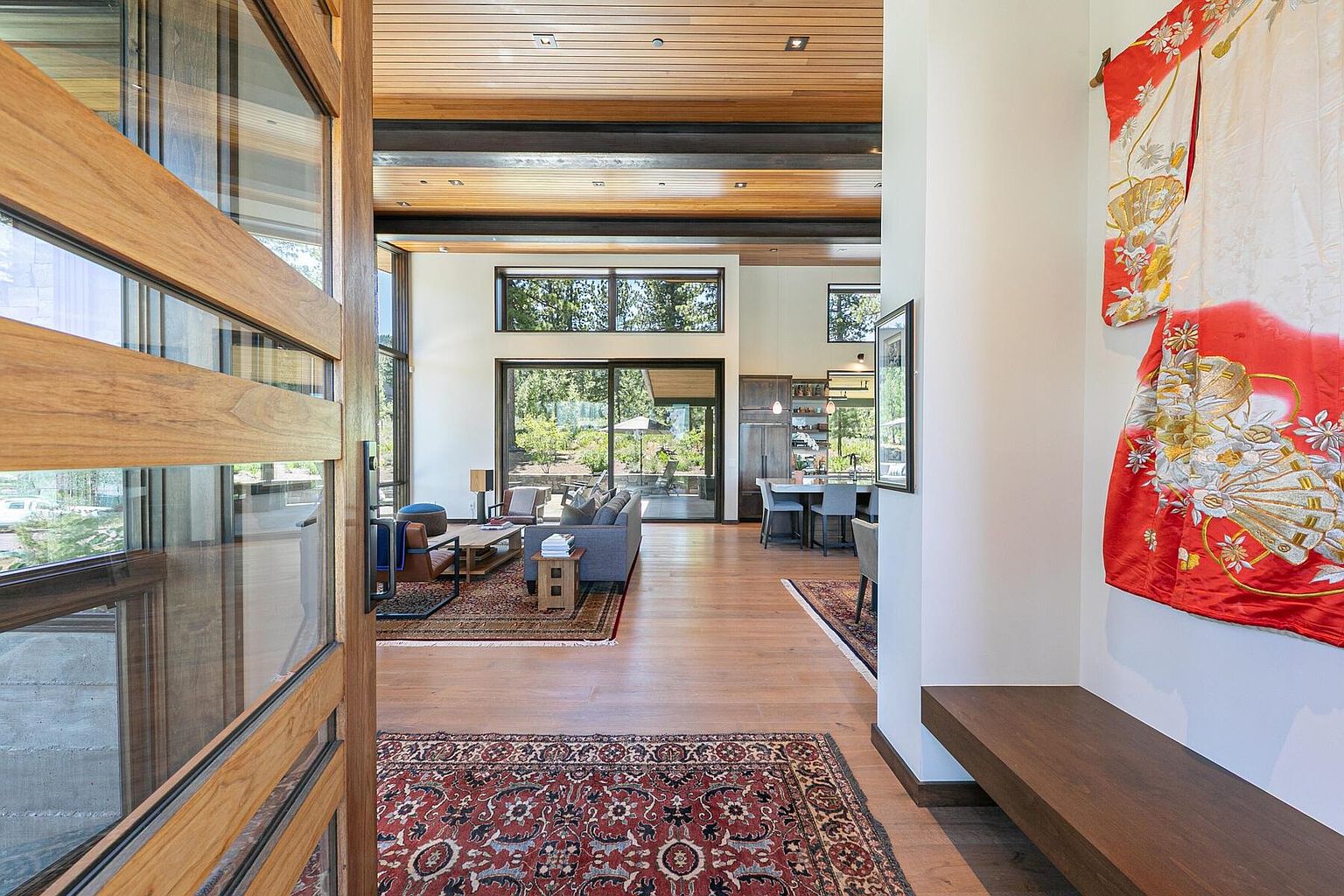 This interior shot showcases a modern hallway leading into a living space. The hallway features hardwood floors, a decorative rug, and a wooden bench against a white wall adorned with a vibrant kimono. The living area beyond includes a gray sofa, a coffee table, and large windows offering natural light and views of the outdoors, creating an inviting and stylish transition between spaces.