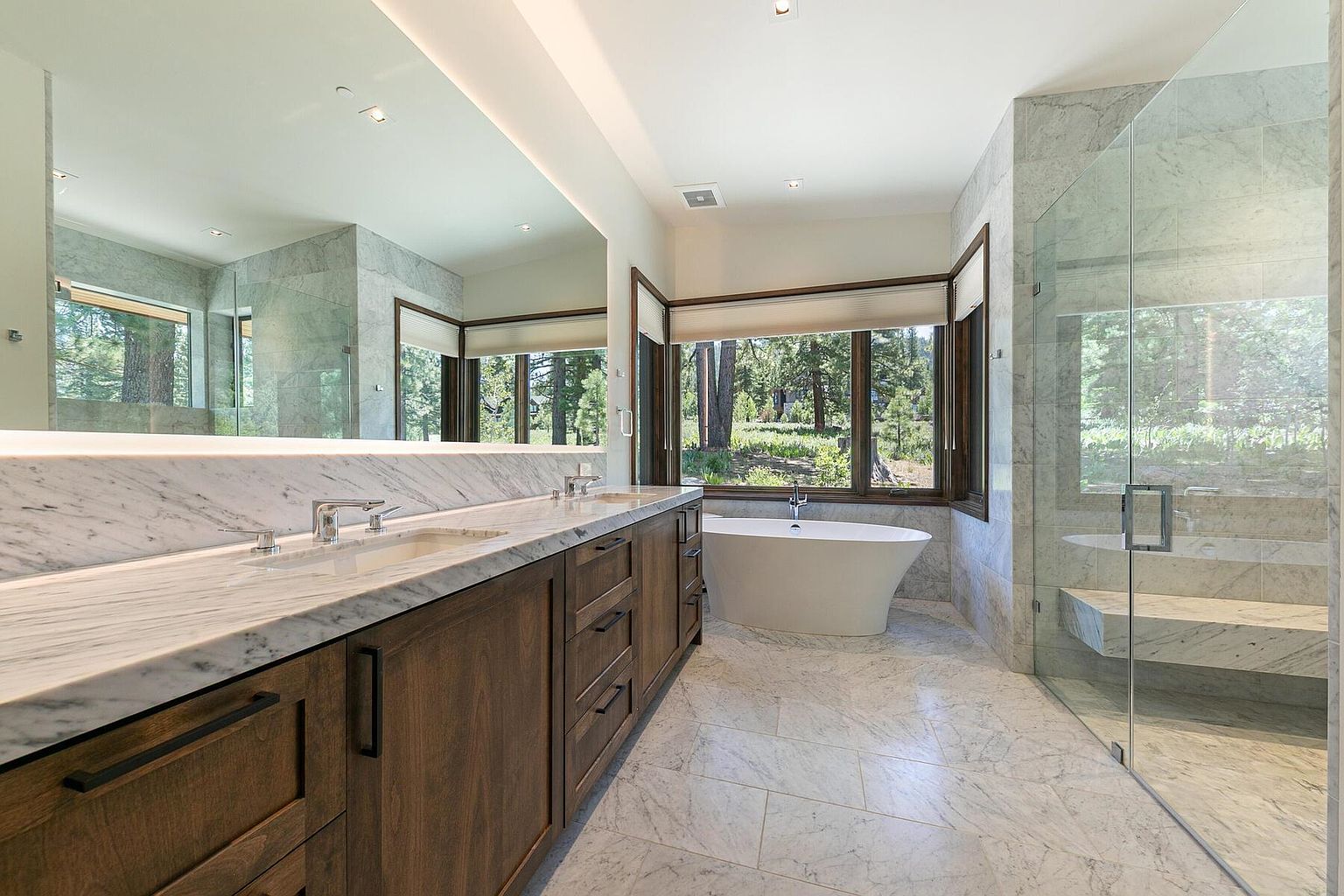 This is a luxurious primary bathroom featuring a double vanity with a marble countertop and dark wood cabinetry. A large mirror spans the length of the vanity, reflecting the natural light from the large window overlooking a wooded area. The bathroom also includes a freestanding soaking tub and a glass-enclosed shower with a built-in bench, all contributing to a spa-like atmosphere.