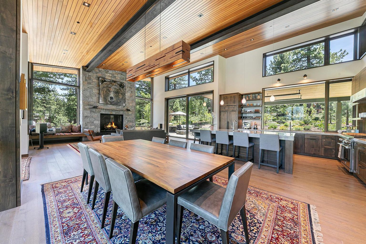 This interior shot showcases a spacious dining area adjacent to an open-concept kitchen and living space. The dining room features a large wooden table with modern chairs, set upon a patterned rug. Natural light floods the area through expansive windows, highlighting the wood-paneled ceiling and stone fireplace, creating a warm and inviting atmosphere.