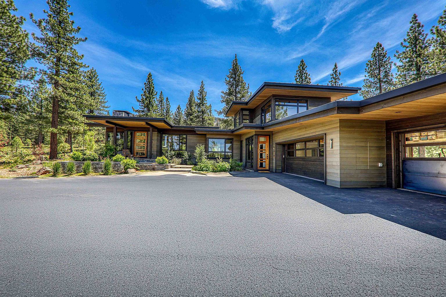This is a front exterior view of a modern, single-story home with a flat roof and a large driveway. The house features a combination of wood and dark-colored siding, with large windows and a prominent entryway. The landscaping is natural and blends with the surrounding trees, creating a serene and private setting.