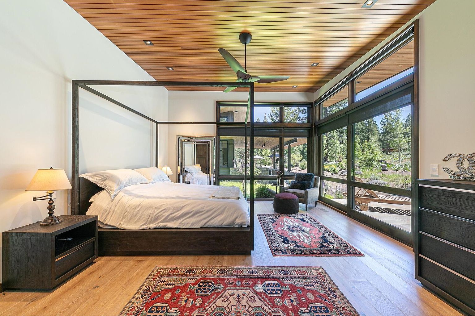 This is a luxurious primary bedroom featuring a four-poster bed with white linens, a dark wood nightstand with a lamp, and a matching dresser. The room boasts a wood-paneled ceiling and large windows offering views of the outdoor landscape. Two decorative rugs add warmth and texture to the hardwood floors.