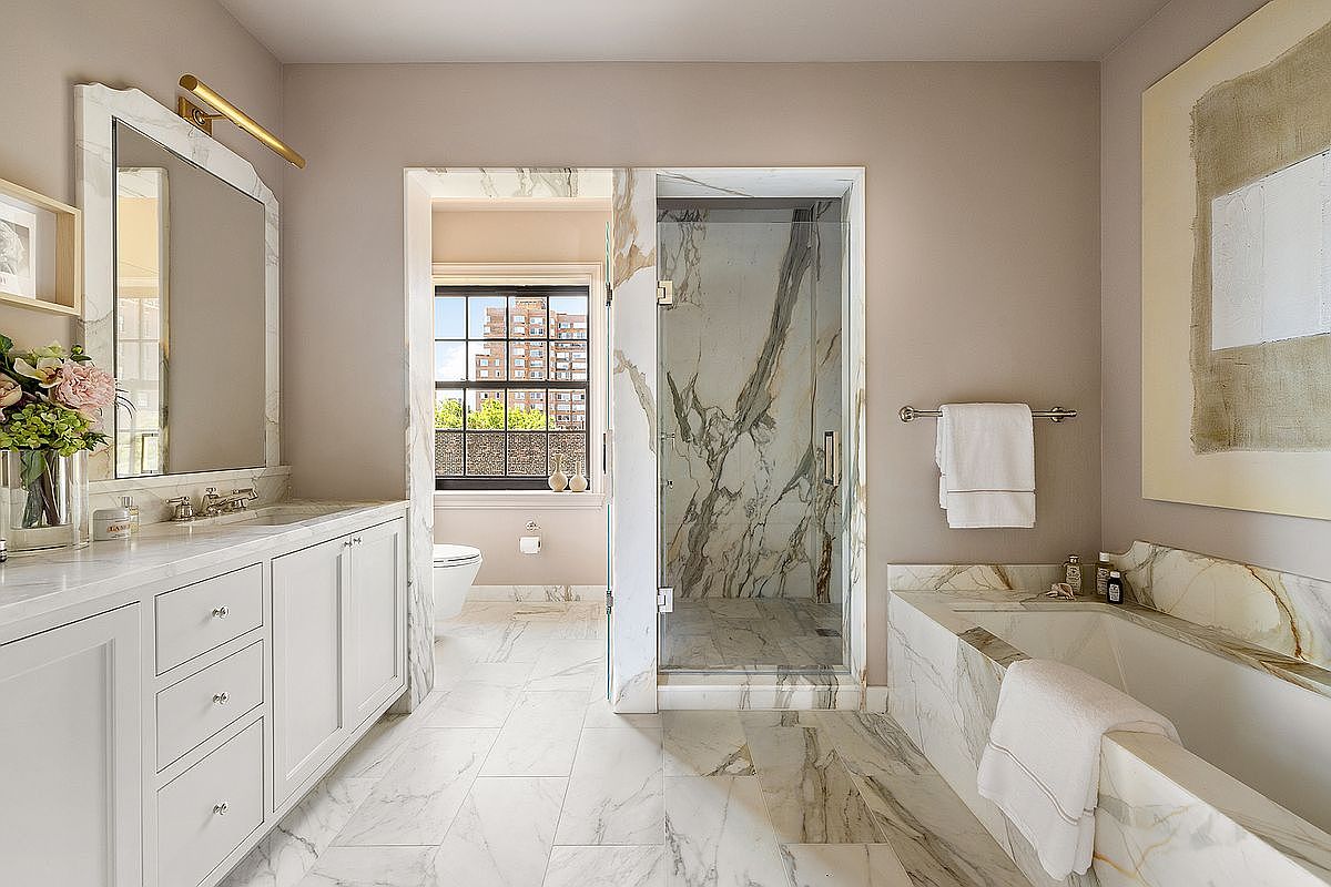 This is a luxurious primary bathroom featuring marble flooring and walls, a white vanity with a marble countertop, and a glass-enclosed shower. A soaking tub is also present, surrounded by marble. The bathroom has a clean and elegant aesthetic, with natural light coming in from a window.