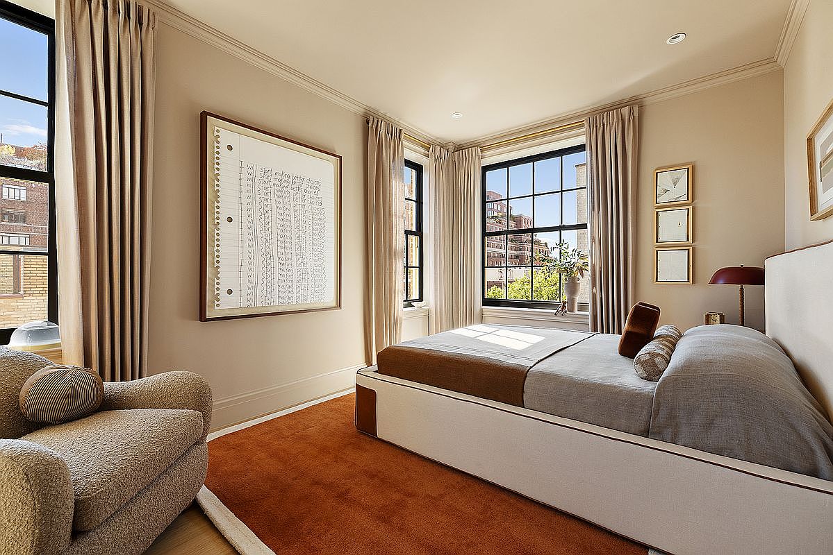 This is a well-lit primary bedroom featuring a large bed with neutral-toned bedding, complemented by an orange rug and beige walls. The room has large windows with views of the city and is decorated with framed artwork, creating a serene and sophisticated atmosphere. A comfortable armchair sits in the corner, adding to the room's inviting appeal.