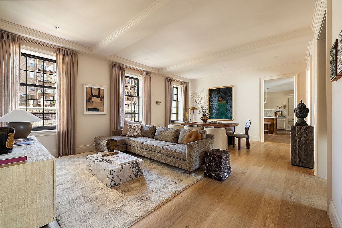 This is a well-lit living room featuring a large sectional sofa, a marble coffee table, and neutral-toned curtains. The room has hardwood floors and a large area rug, creating a cozy and inviting atmosphere. Artwork adorns the walls, and there is a glimpse into another room through an open doorway, suggesting an open floor plan.