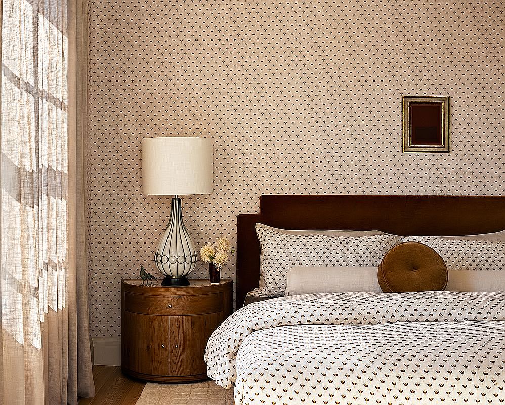 This is a cozy primary bedroom featuring a patterned wallpaper and bedding. A wooden nightstand with a lamp sits beside the bed, and a framed picture hangs on the wall above the headboard. The room has a warm and inviting atmosphere, enhanced by natural light filtering through the curtains.