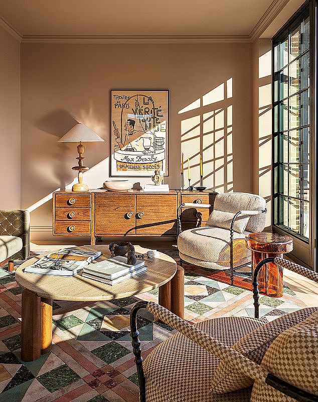 This interior shot showcases a stylish living room with a vintage-inspired aesthetic. The room features a wooden sideboard with a lamp and artwork above, a round coffee table with books, and patterned armchairs. Natural light streams through a large window, highlighting the unique geometric tile flooring and creating a warm, inviting atmosphere.