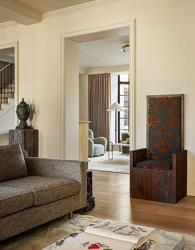 This interior shot showcases a well-appointed living room with a blend of modern and classic elements. A comfortable brown sofa is positioned near a unique, high-backed chair with patterned upholstery. The room opens into another space through an arched doorway, suggesting an open floor plan and creating a sense of spaciousness.