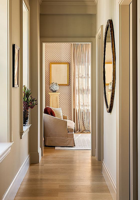 This is an interior shot of a hallway leading into a living space. The hallway features hardwood floors, neutral-colored walls, and decorative trim. A unique oval mirror hangs on the wall, and the living space beyond is decorated with patterned wallpaper, a framed picture, and a comfortable armchair, creating an inviting and stylish transition between spaces.