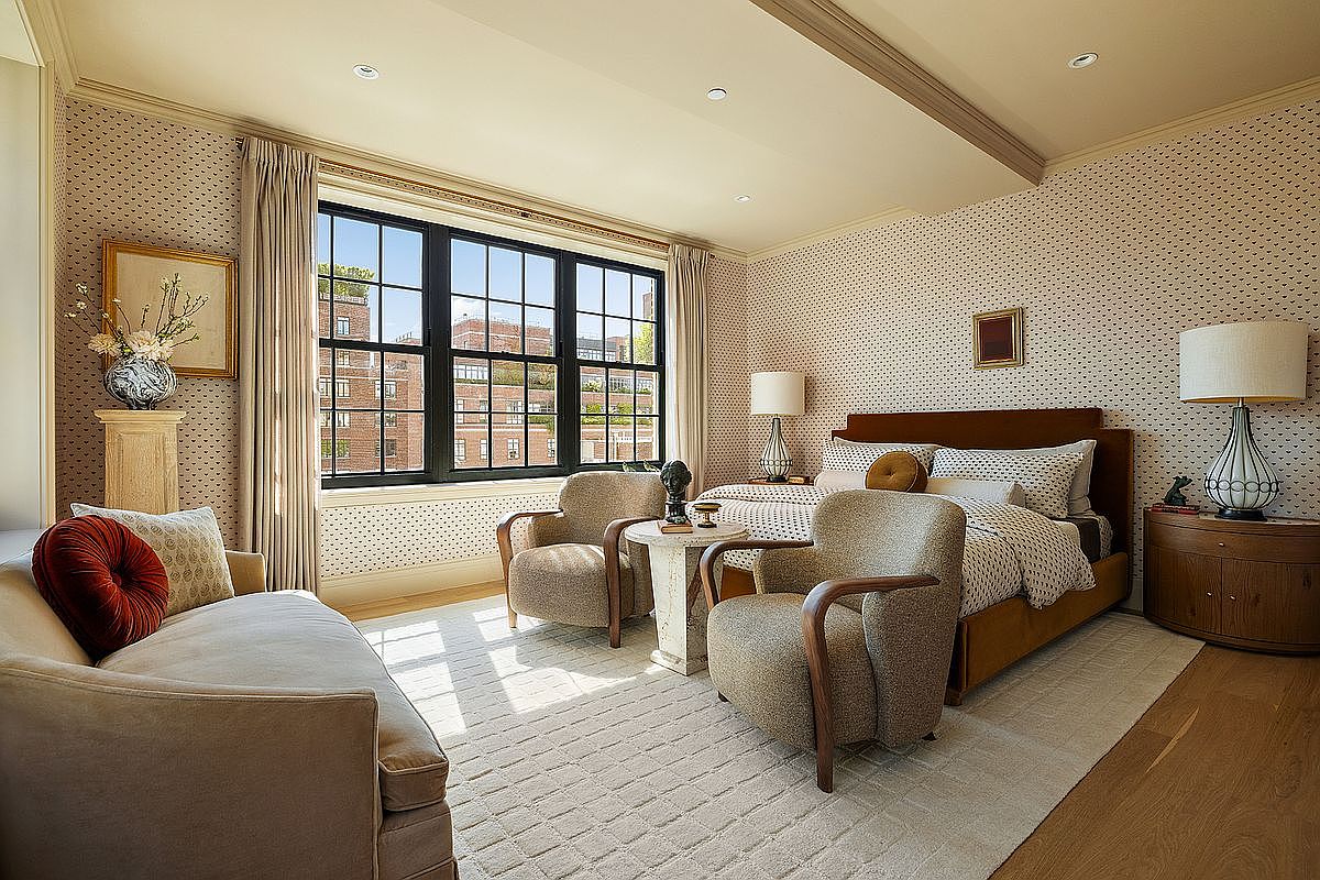 This is a well-lit primary bedroom featuring a large window with black frames offering a city view. The room is furnished with a bed, two armchairs, a small table between them, and a sofa. The walls are covered in patterned wallpaper, and a light-colored rug covers most of the wooden floor, creating a cozy and inviting atmosphere.
