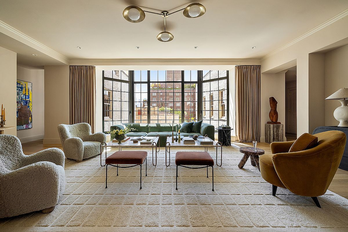This is a spacious living room featuring a large window with black frames offering city views. The room is furnished with a green sofa, two armchairs, and two ottomans, all arranged on a patterned rug. The color palette is neutral with pops of green and brown, creating a sophisticated and inviting atmosphere.
