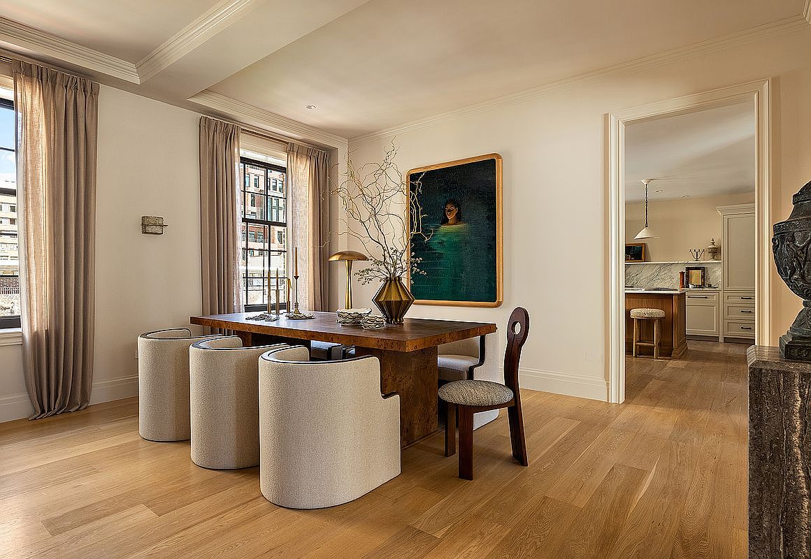 This is an interior shot of a dining room featuring a wooden table surrounded by modern, upholstered chairs and a unique wooden chair. A large painting hangs on the wall, complemented by a decorative vase and a lamp on the table. The room has hardwood floors and a doorway leading to another room, possibly a kitchen, creating an open and inviting atmosphere.