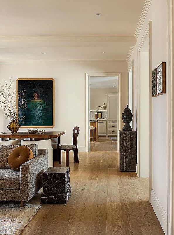This interior shot showcases a stylish living room that seamlessly blends into the dining area and offers a glimpse into the kitchen. The space features hardwood flooring, neutral-toned walls, and sophisticated furnishings, including a comfortable sofa with a round accent pillow and a unique marble side table. A large painting adds a touch of artistry, while the open doorway invites exploration of the home's layout.