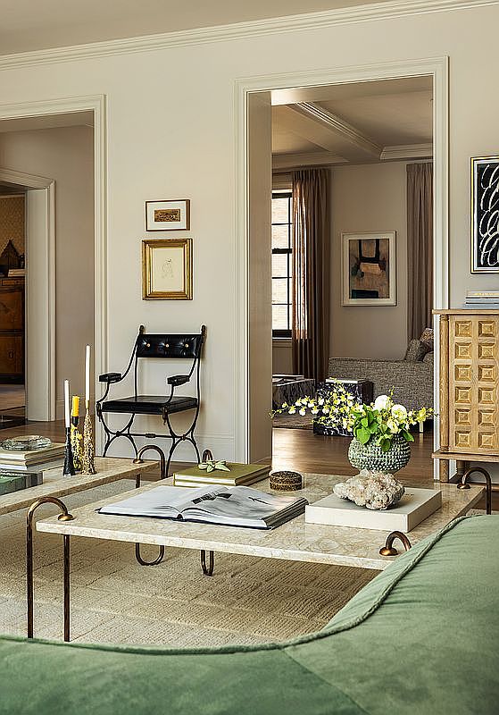 This is an interior shot of a living room featuring a stylish arrangement of furniture and decor. The room includes a marble-topped coffee table with books and decorative items, a black chair, and a glimpse into an adjacent room with curtains and artwork. The overall impression is one of sophisticated elegance and comfortable living.