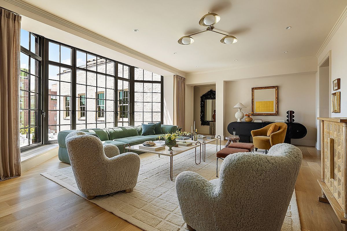 This is a bright and airy living room featuring a large window with black frames, offering ample natural light. The room is furnished with a green sectional sofa, two textured armchairs, and a marble-topped coffee table, all set on a patterned area rug. A modern chandelier hangs from the ceiling, and a dark sideboard with decorative items adds a touch of elegance to the space.