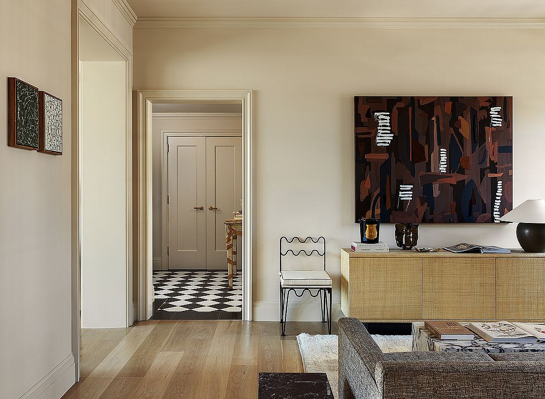 This interior shot showcases a stylish living room with hardwood floors and neutral-toned walls. A large abstract painting hangs above a woven console table, complemented by a unique chair and a glimpse of a black and white checkered floor through a doorway. The room exudes a sophisticated and artistic ambiance.