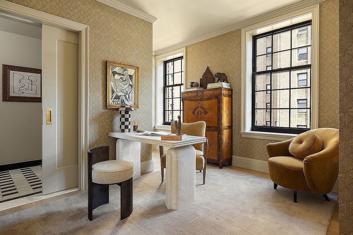 This is an elegant home office featuring patterned wallpaper, two windows with black frames, and a unique white desk with sculptural legs. A vintage wooden cabinet adds character, while a mustard-colored armchair provides a comfortable seating area. The room exudes a sophisticated and artistic ambiance.