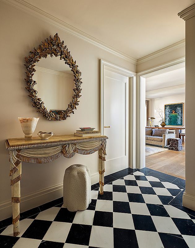 This interior shot showcases a classically styled hallway with a black and white checkered tile floor. A decorative console table with an ornate mirror above it adds a touch of elegance. The hallway leads into a living area, visible through an open doorway, suggesting a seamless flow between spaces.