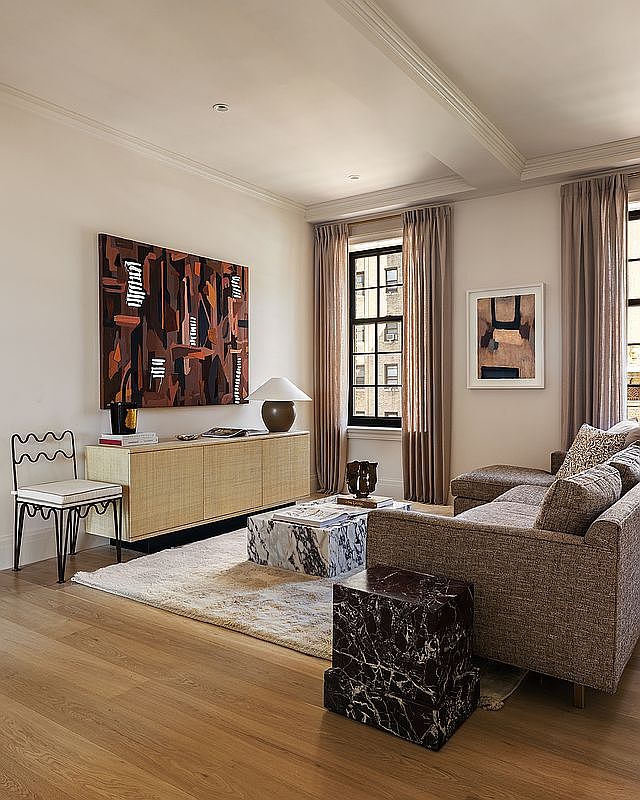 This is an interior shot of a living room featuring a neutral color palette with hardwood floors and a plush rug. The room is furnished with a brown sofa, marble coffee table, and a console table with artwork above. Natural light streams in through the windows, creating a warm and inviting atmosphere.