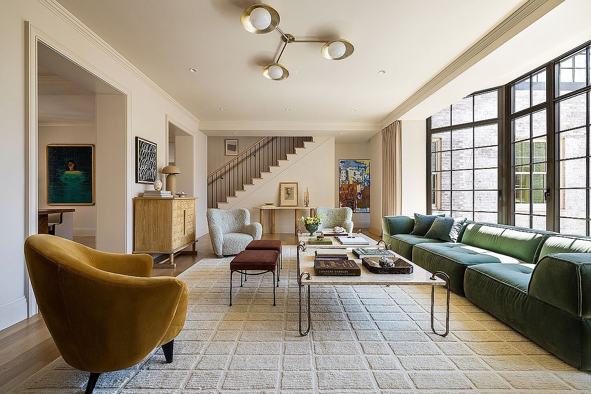 This is a well-lit living room featuring a large window with black frames, a green sectional sofa, and a light-colored rug with a grid pattern. The room also includes a coffee table with books and decorative items, a yellow armchair, and a staircase in the background. The overall impression is one of modern elegance and comfort.