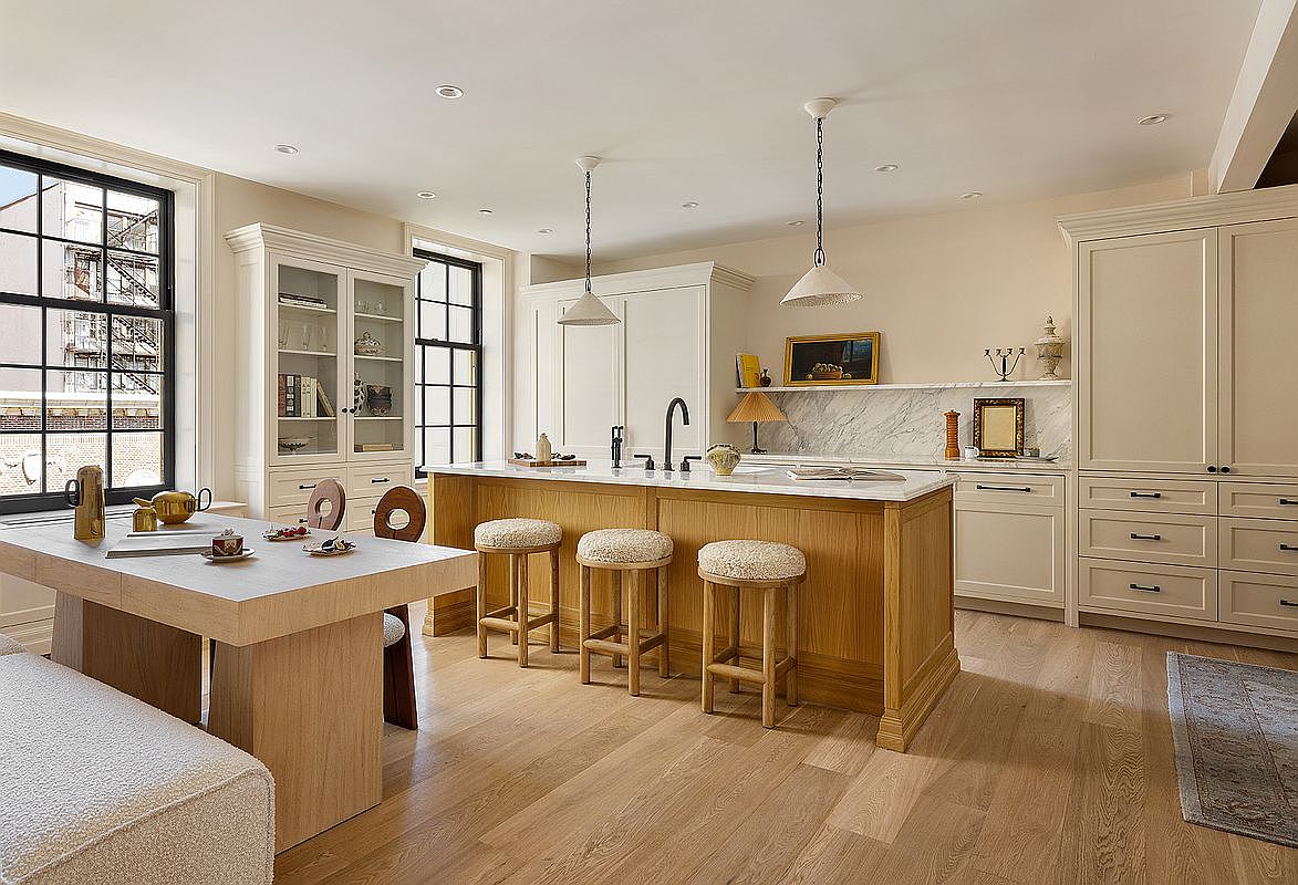 This is a bright and airy kitchen featuring a large wooden island with a white countertop and seating for three. The kitchen also includes custom cabinetry, a marble backsplash, and modern pendant lighting. A dining table with a bench and chairs sits adjacent to the kitchen area, creating an open and inviting space.