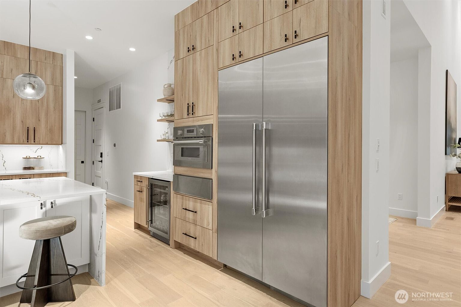 This is a bright and modern kitchen featuring light wood cabinetry and stainless steel appliances, including a large refrigerator. The kitchen island has a white countertop and a stool, and there are open shelves displaying decorative items. The overall impression is clean, stylish, and functional, perfect for a modern home.