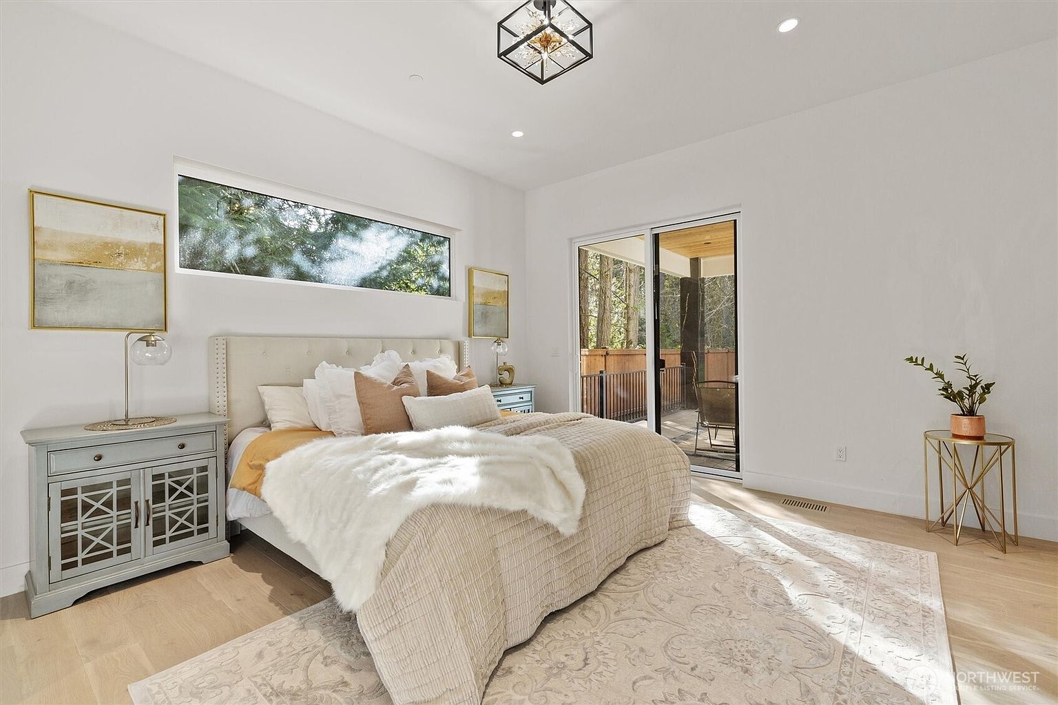 This is a bright and airy primary bedroom featuring a neutral color palette. The room includes a large bed with a plush headboard, multiple pillows, and a soft throw blanket. Natural light floods the space through a horizontal window and a sliding glass door leading to an outdoor area, complemented by stylish side tables and artwork.