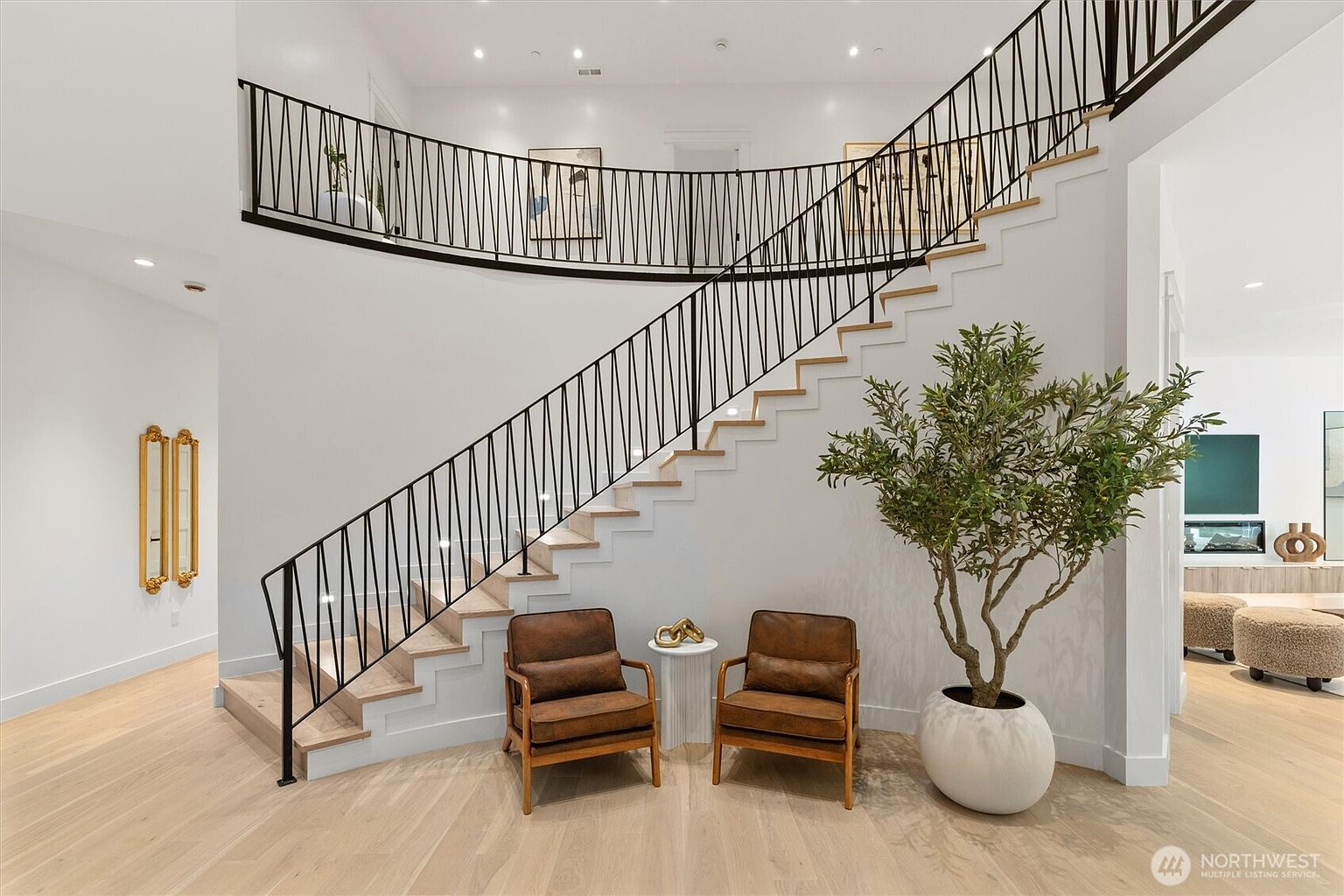This interior shot showcases a grand hallway with a modern staircase featuring a black railing and light wood steps. Two brown leather chairs flank a small white table near the base of the stairs, complemented by a large potted tree. The space is well-lit, highlighting the clean lines and minimalist design, creating an inviting and sophisticated atmosphere.