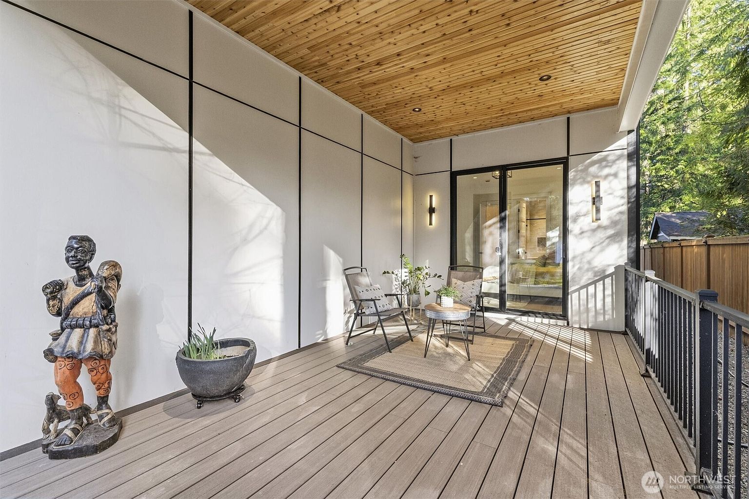 This image showcases a stylish outdoor patio or deck area. The space features modern design elements, including a wooden ceiling, sleek wall paneling with black accents, and contemporary outdoor furniture. The deck is furnished with chairs, a small table, and a decorative rug, creating an inviting and comfortable outdoor living space.